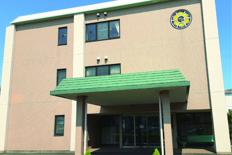 緑色の庇と時計が特徴の介護施設の建物外観。レンガ造りの3階建て。